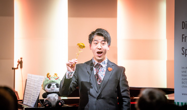 Im Zentrum des Bildes steht ein freundlich lächelnder Mann im Anzug. Er singt und  hält eine goldene Rose halb hoch. Ein Panda Stofftier sitzt auf einem und ein Flügel hinter dem singenden Mann. Im Hintergrund gedimmtes Licht. 