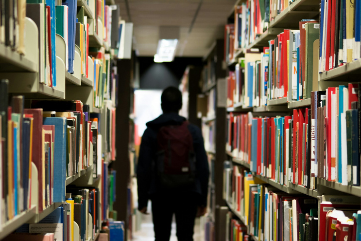 Im Gang einer modernen Bibliothek steht im Fluchtpunkt zweier Bücherregale eine Person die den Betrachtern den Rücken zuwandt.