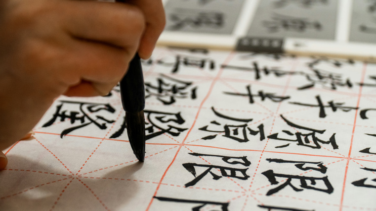 Im Bild sieht man eine Hand die einen Kalligraphie Pinsel hält. Pinsel und Hand sind gerichtet auf ein Kalligraphiepapier und schreiben chinesiche Schriftzeichen die in großen, dicken Strichen auf das Papier geschrieben werden.