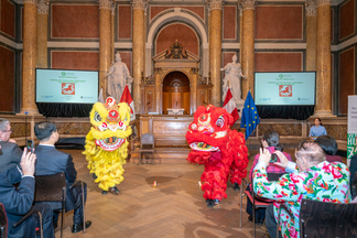 Im großen Festsaal der Universität Wien tanzen zwei Löwen und begrüßen das chinesische Neujahr. Im Vordergrund dreht sich ein Mann nach außen um die tanzenden Löwen zu fotographieren.